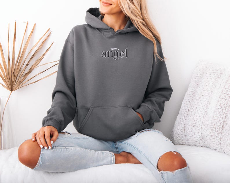 May include: A charcoal gray hoodie with the word "angel" embroidered on the chest. The hoodie has a front pocket and a hood. The model is wearing light wash ripped jeans. The background includes a dried palm leaf and a white pillow.