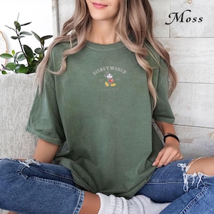 Puede incluir: Una camiseta verde musgo con el texto "DISNEY WORLD FLORIDA" y un gr&aacute;fico de Mickey Mouse. La camiseta tiene mangas cortas y cuello redondo. La palabra "Moss" est&aacute; escrita en cursiva en la esquina superior derecha.