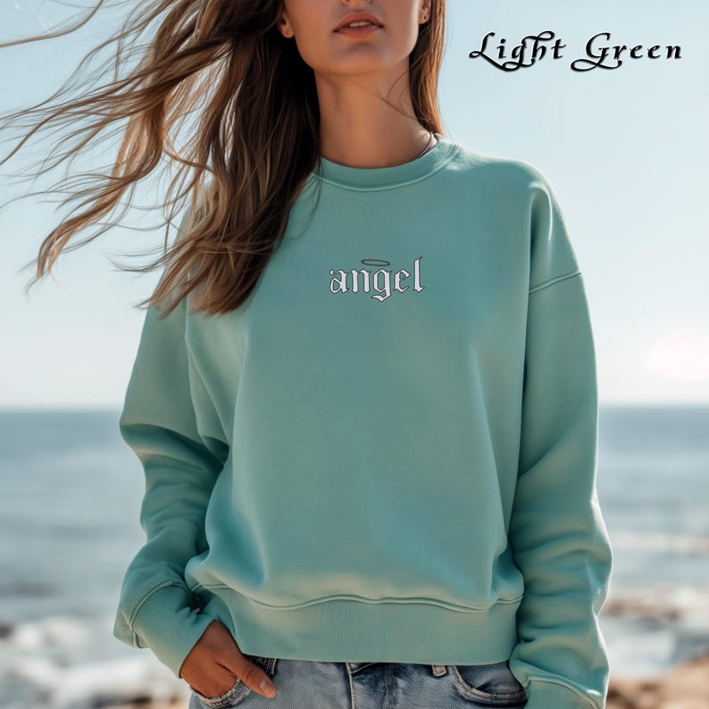 Pode incluir: Uma camisola de cor verde claro com a palavra "angel" bordada na frente em branco com uma aur&eacute;ola acima da palavra.