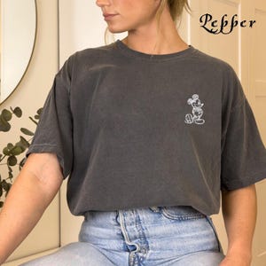 May include: A dark grey t-shirt featuring a white embroidered Mickey Mouse graphic. The t-shirt is being worn by a person with long brown hair and is paired with light blue jeans.