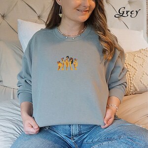 Op de afbeelding: Een grijs sweatshirt met een afbeelding van vijf vrouwen die samen staan. De vrouwen hebben allemaal verschillende huidtinten en verschillende kapsels. Het sweatshirt is een ronde hals met lange mouwen.