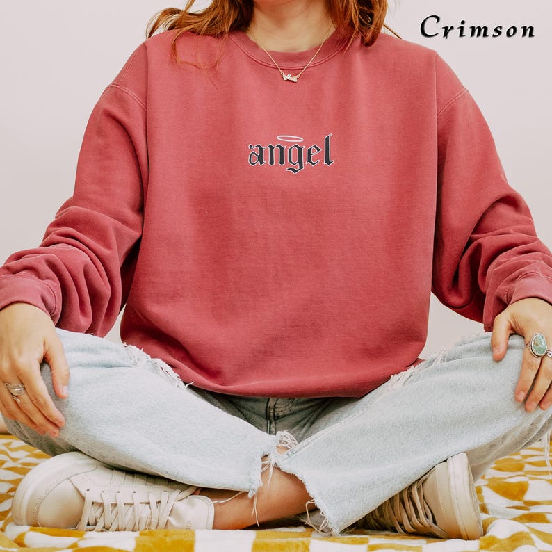 Pode incluir: Uma camisa de moletom vermelha carmim com a palavra "angel" em letras g&oacute;ticas pretas com uma aur&eacute;ola acima.