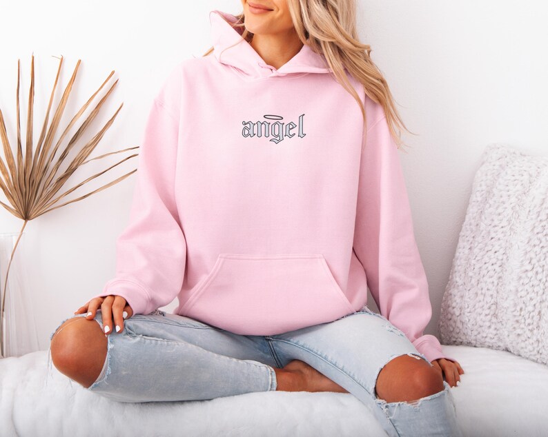 May include: A light pink hoodie with a front pocket and a hood. The word "angel" is embroidered in black script on the chest, with a halo above the word. The hoodie is made of a soft material.
