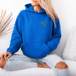 May include: A royal blue hoodie with a kangaroo pocket and drawstring hood. The hoodie features a small embroidered design of a green frog holding a drink. The model is wearing light blue jeans with ripped knees.