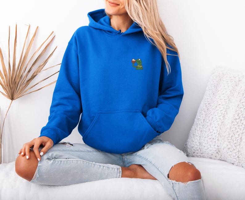 May include: A royal blue hoodie with a kangaroo pocket and drawstring hood. The hoodie features a small embroidered design of a green frog holding a drink. The model is wearing light blue jeans with ripped knees.