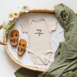 May include: A beige baby onesie with the text "baby anderson" and a heart symbol, placed on a white cloth inside a woven basket. Two brown suede baby shoes and a green muslin blanket are also in the basket. White flowers are on the left.