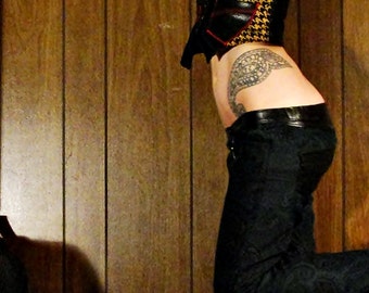 May include: A woman wearing a black and yellow patterned top with a black bra underneath. She is wearing black jeans and has a tattoo on her hip and arm. The background is a wood panel wall.
