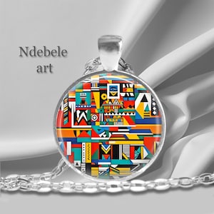 May include: A silver chain necklace with a round pendant featuring a colorful geometric pattern inspired by Ndebele art. The pendant is made of glass and has a silver rim.