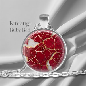 May include: A silver necklace with a round pendant featuring a red and gold kintsugi design. The pendant has a white background with gold cracks and red filling. The text "Kintsugi Ruby Red" is visible above the pendant.