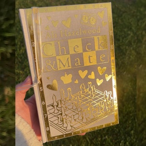Może przedstawiać: Książka zatytułowana "Check & Mate" autorstwa Ali Hazelwood. Okładka jest beżowa ze złotymi akcentami, w tym sercami, figurami szachowymi i wzorem szachownicy. Książka jest trzymana w dłoni.