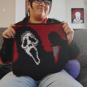 Puede incluir: Un cojín tejido a mano con el diseño de la máscara de Ghostface de la serie de películas de terror Scream. El cojín tiene un fondo negro con un borde rojo y un diseño de máscara blanca y gris. También se ve un cuchillo tejido.