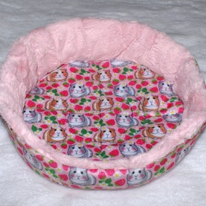 May include: A pink and white round pet bed with a soft, fluffy pink rim. The bed is lined with a pink fabric featuring a pattern of small guinea pigs and strawberries.