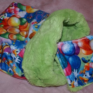 May include: A green plush fabric baby blanket with a colourful fabric insert featuring a cartoon character and balloons. The insert is attached to the blanket with a green trim.