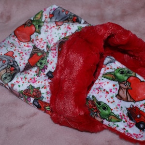 May include: A red and white fabric baby blanket with a Valentine's Day theme. The blanket features a repeating pattern of small hearts and cartoon characters holding hearts. One side of the blanket is red fleece.