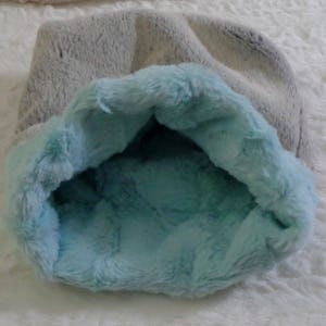 May include: A grey pet bed with a light blue interior, made from a soft, plush material. The bed is designed to provide a cosy and comfortable space for a small animal. The fluffy texture is visible.