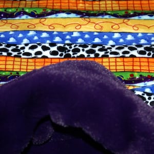May include: A close-up of a plush, deep purple fabric draped over a colourful patterned fabric. The patterned fabric features horizontal stripes in orange, yellow, blue, and white with various designs, including clouds and cow spots.