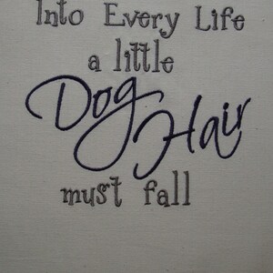 May include: A beige fabric with the text "Into Every Life a little Dog Hair must fall" embroidered in black and gray thread.