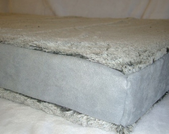 May include: A rectangular pet bed with a plush, faux fur top in shades of gray and white. The sides are a smooth, light gray material. The bed is designed for comfort and support.