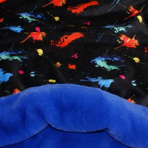 May include: A black and blue fleece blanket with a colourful paint splatter pattern. The blanket is soft and cosy, perfect for snuggling up on the sofa.