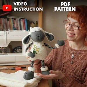 May include: A handcrafted sheep soft toy with a white, fleecy body, grey face, ears, and feet, and striped arms and legs. The doll features a small bunch of yellow flowers on its chest. The image also includes the text "VIDEO INSTRUCTION" and "PDF PATTERN".