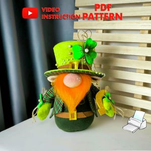 Puede incluir: Un muñeco de duende de fieltro verde con un sombrero verde, barba naranja y una olla de oro. El muñeco sostiene un trébol de cuatro hojas en su mano. El muñeco está sentado sobre una superficie blanca. La imagen incluye el texto "VIDEO INSTRUCTION PATTERN" y "PDF PATTERN".