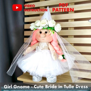 May include: A white tulle dress with a white lace bodice and a white flower crown. The doll has pink hair and a white hat. The doll is standing on a wooden shelf. "Girl Gnome - Cute Bride in Tulle Dress" is written below the doll.