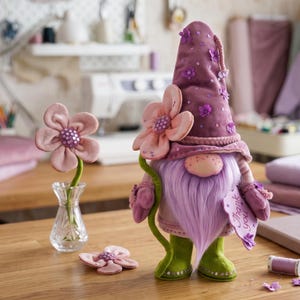 Può includere: Una statuetta di gnomo in peluche che indossa un alto cappello color malva decorato con paillettes viola e piccoli fiori di stoffa. Lo gnomo ha una lunga barba color lavanda, stivali verdi e tiene in mano un fiore rosa e un cartellino con il testo Thank you.
