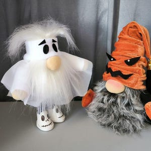 May include: Two Halloween gnomes, one dressed as a ghost with a white fuzzy beard and a white sheet, and the other dressed as a pumpkin with an orange velvet hat and a gray fuzzy beard.