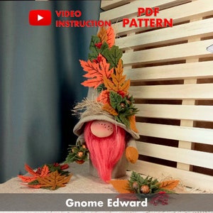 Può includere: Un gnomo di feltro con la barba rosa e un cappello decorato con foglie autunnali. Lo gnomo tiene un mazzo di foglie e ghiande di feltro. Il testo "Gnome Edward" è sotto lo gnomo.