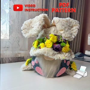 Op de afbeelding: Een pluche paasbunny-gnome met een wit bontlichaam, blauwe spijkerbroek, roze voeten en een gele bloemenkrans. De gnome houdt een geel kuiken in zijn hand. De afbeelding bevat de tekst "VIDEO INSTRUCTION" en "PDF PATTERN".