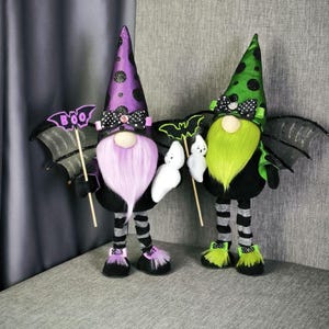 Pode incluir: Duas decorações de gnomos de Halloween. Um gnomo tem um chapéu roxo com bolinhas pretas, uma barba rosa e uma placa que diz "BOO". O outro gnomo tem um chapéu verde com bolinhas pretas e uma barba verde. Ambos têm asas de morcego e seguram fantasmas.