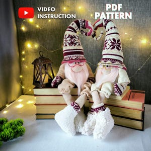 Peut inclure: Deux poupées gnomes faites à la main avec de longues barbes blanches, portant des lunettes et des chapeaux à motifs. Les chapeaux sont de couleur crème avec des motifs de flocons de neige bordeaux. Les poupées sont assises sur une pile de livres. Le texte "VIDEO INSTRUCTION" et "PDF PATTERN" est inclus.