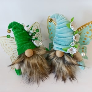 May include: Two gnomes with butterfly wings. One gnome has a green velvet hat and the other has a blue velvet hat. Both gnomes have brown fur beards and white flowers on their hats.