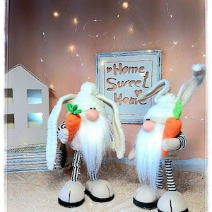 Könnte beinhalten: Zwei weiße Plüschgnome mit Hasenohren und gestreiften Hosen, die Karotten halten. Die Gnome stehen auf einer weißen gehäkelten Oberfläche. Ein Schild hinter den Gnomen lautet "Home Sweet Home".
