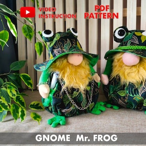 May include: Two green frog gnome plush toys with large eyes, wearing green hats with leaf patterns and holding a chain. The gnomes have long yellow beards and are standing on a green surface. The text "GNOME Mr. FROG" is at the bottom of the image.