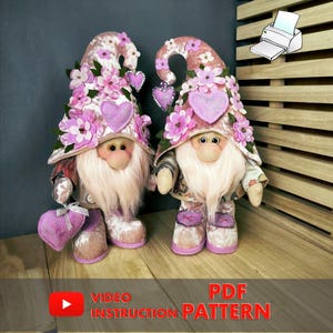 Può includere: Due gnomi di peluche con lunghe barbe bianche, che indossano cappelli rosa adornati con fiori e dettagli a forma di cuore. Hanno stivali rosa abbinati e tengono cuscini a forma di cuore. È presente anche il testo "PDF INSTRUCTION PATTERN".