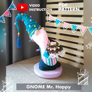 May include: A blue and white gnome holding a pink and brown cake with the text "GNOME Mr. Happy" on a pink base. The gnome is wearing a blue hat with white pom-poms and a blue and white striped shirt. The background is a wooden slat wall with a pink and blue banner.