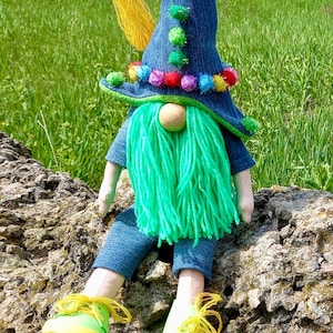 Może przedstawiać: Zielono-niebieska lalka gnoma w dżinsowym kapeluszu z kolorowymi pomponami i zieloną brodą z włóczki. Gnom siedzi na skale z zieloną trawą w tle.