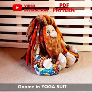 May include: A plush gnome doll wearing a colorful yoga outfit, sitting in a meditation pose. The gnome has long, brown and orange dreadlocks, a brown beard, and blue eyes. The doll is sitting on a brown surface with a wooden slatted background. The text "Gnome in YOGA SUIT" is at the bottom of the image.
