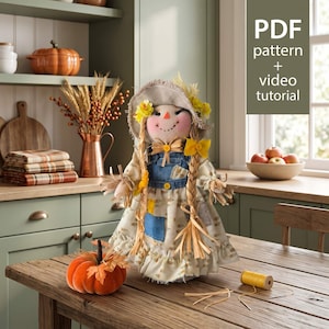 May include: A fabric scarecrow doll wearing a straw hat, a blue denim overall dress, and a patterned beige dress with yellow accents. The doll has braided yarn hair and is positioned next to a small orange fabric pumpkin and a spool of yellow thread.