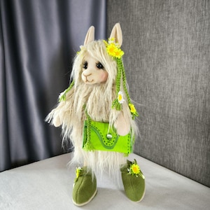 Puede incluir: Una muñeca de llama hecha a mano con un cuerpo color crema y pelaje largo y lanudo. La llama lleva un bolso verde, zapatos y un adorno floral con flores amarillas. La muñeca tiene largas pestañas y una expresión dulce.