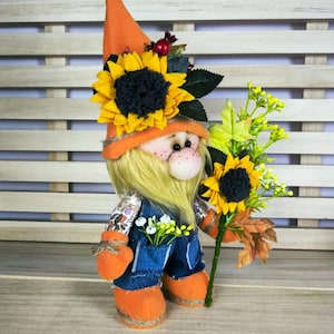 Può includere: Una bambola di gnomo fatta a mano con cappello e stivali arancioni, tuta blu e lunga barba gialla. Lo gnomo è decorato con girasoli e tiene in mano un mazzo di fiori e foglie. La bambola è ambientata su uno sfondo di legno chiaro.