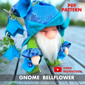 May include: A blue and green gnome doll with a blue sequined hat and a white beard. The gnome is wearing a green shirt and blue sequined sleeves. The gnome is holding a blue flower in its hand. The image includes the text "PDF PATTERN", "VIDEO INSTRUCTION", and "GNOME BELLFLOWER".
