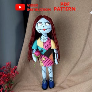 May include: A handmade doll with red hair, blue and green patchwork clothing, and a stitched-on face. The doll is holding a pink flower and is standing on a brown surface. The image includes the text "VIDEO INSTRUCTION" and "PDF PATTERN".
