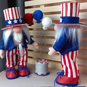 Puede incluir: Dos gnomos patrióticos que llevan sombreros y atuendos rojo, blanco y azul. Están sosteniendo globos y de pie frente a un fondo de madera.