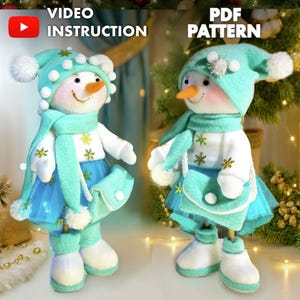 以下が含まれることがあります： 2体のぬいぐるみ雪だるま。各雪だるまは、ターコイズの帽子、スカーフ、青いスカートの衣装を着ています。雪だるまは、人参の鼻、笑顔の顔、白いブーツをしています。画像には「VIDEO INSTRUCTION」と「PDF PATTERN」の文字が含まれています。
