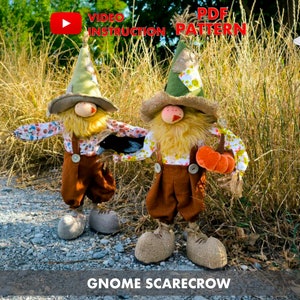 Puede incluir: Dos espantapájaros enanos con pantalones marrones, camisas blancas con estampados florales y sombreros verdes. El gnomo de la derecha sostiene calabazas naranjas. Los gnomos están de pie en un camino de grava frente a hierba alta.