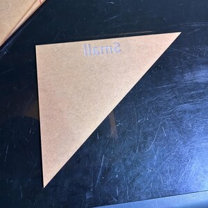 K&ouml;nnte beinhalten: Eine transparente, dreieckige Schablone mit dem Wort "Small" auf der Oberfl&auml;che. Die Schablone besteht aus einem durchscheinenden, hellbraunen Material. Die Schablone liegt auf einer dunklen Oberfl&auml;che.
