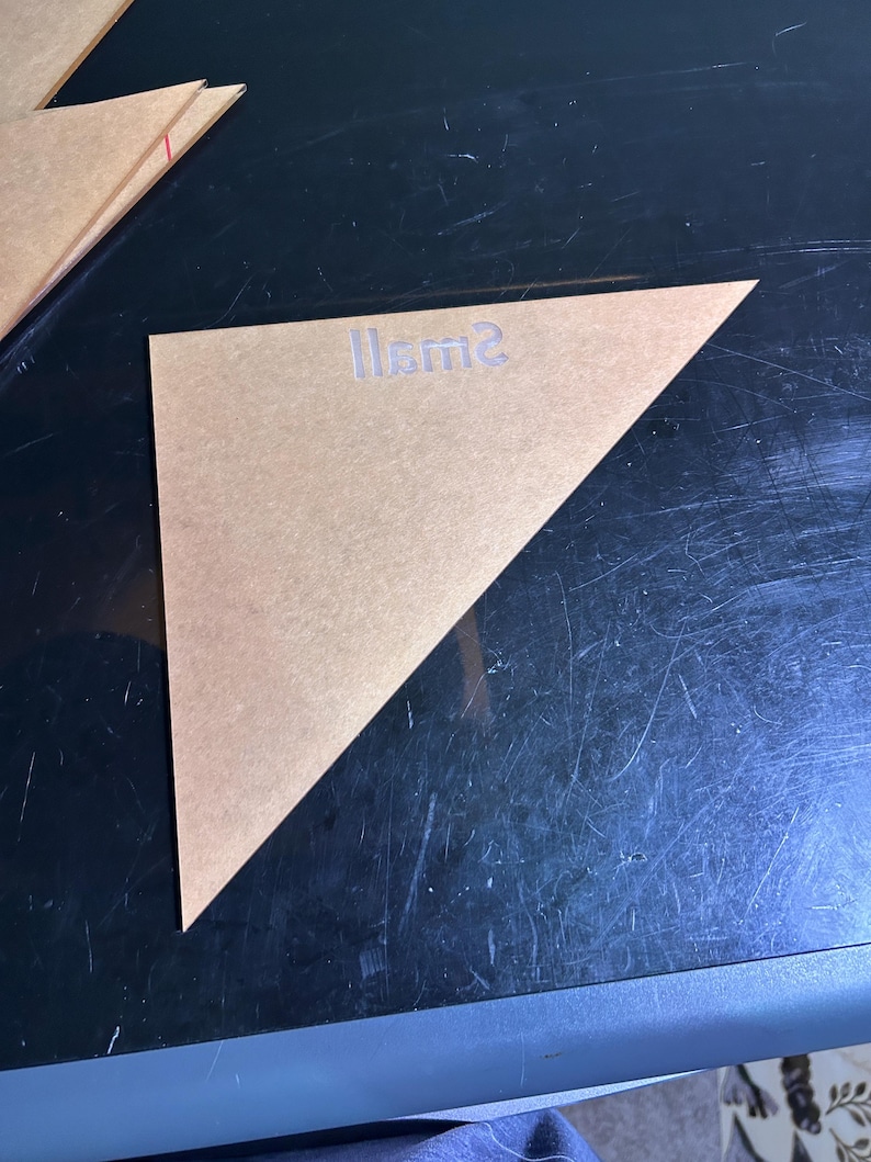 K&ouml;nnte beinhalten: Eine transparente, dreieckige Schablone mit dem Wort "Small" auf der Oberfl&auml;che. Die Schablone besteht aus einem durchscheinenden, hellbraunen Material. Die Schablone liegt auf einer dunklen Oberfl&auml;che.