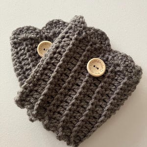 Può includere: Due scaldino per tazze in crochet grigio con bottoni in legno. I bottoni hanno la scritta "Handmade with love".